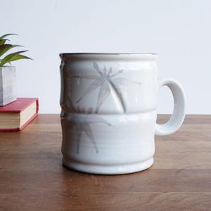 May include: White ceramic mug with a gray floral design. The mug has a rounded handle and a slightly flared rim.