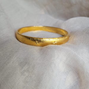 Puede incluir: Un brazalete dorado con superficie texturizada y grabados en forma de estrella. El brazalete es circular y parece ser de metal. El fondo es una tela blanca y suave.