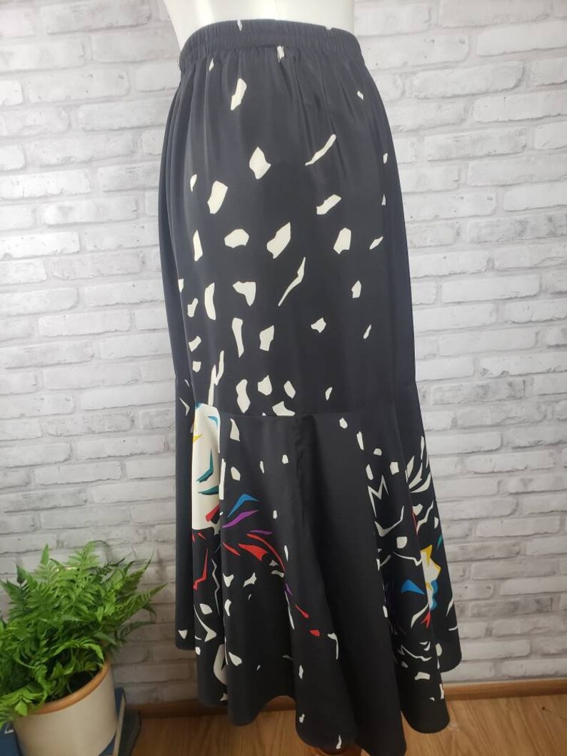 May include: A black midi skirt with a white and colorful abstract print. The skirt has a gathered hem and an elastic waistband.