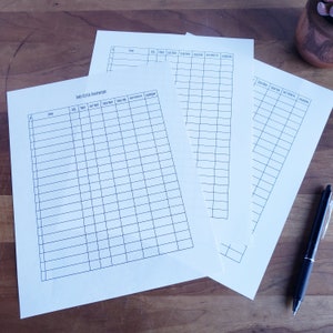May include: Several cream-colored inventory sheets with black grid lines and headings, including "Item," "Cost," and "Sold Date." A black pen rests on the wooden surface next to the sheets. The text "Etsy Listed Inventory" is visible on one sheet.