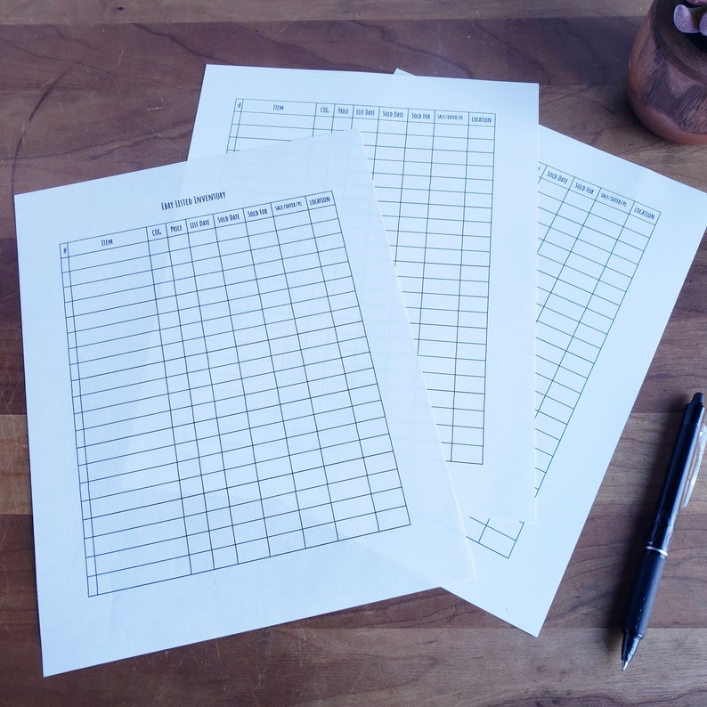 May include: Several cream-colored inventory sheets with black grid lines and headings, including "Item," "Cost," and "Sold Date." A black pen rests on the wooden surface next to the sheets. The text "Etsy Listed Inventory" is visible on one sheet.