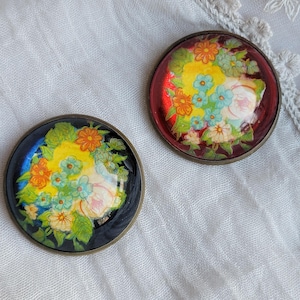 Op de afbeelding: Twee ronde broches met bloemmotieven. De ene broche heeft een zwarte achtergrond, de andere een rode achtergrond. Beide broches hebben een kleurrijk bloemenontwerp met gele, oranje, blauwe en witte bloemen. De broches hebben een bronzen metalen rand.