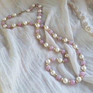 May include: A beaded necklace featuring alternating round white pearls and translucent light pink beads, separated by small gold-toned metal spacers. The necklace includes a gold-toned metal clasp closure at the end of the strand of beads.