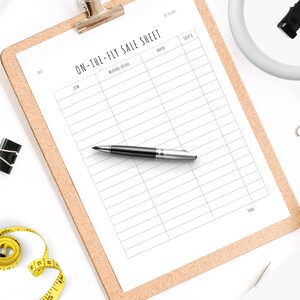 May include: A white clipboard with a cork backing, labeled "On-the-Fly Sale Sheet". The clipboard has columns for date, item, measurements/details, buyer, sold for, and total. A pen is lying on top of the clipboard.