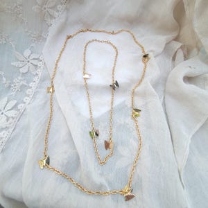 May include: A gold-coloured necklace featuring several butterfly charms. The necklace is displayed on white fabric. The butterflies have a metallic finish with touches of green, gold, and brown. The chain is delicate and has a link design.