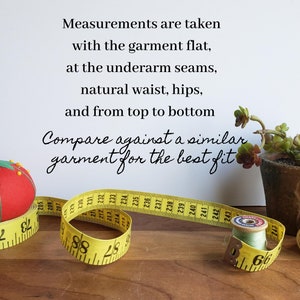 May include: Yellow measuring tape with black markings, a red pincushion in the shape of a tomato, and a spool of thread. The tape reads "Measurements are taken with the garment flat, at the underarm seams, natural waist, hips, and from top to bottom. Compare against a similar garment for the best fit."