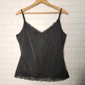 May include: A black camisole featuring a V-neckline with delicate lace trim and matching lace detailing along the bottom hem. The garment has thin, adjustable spaghetti straps and a smooth, solid-colored fabric construction throughout the body.