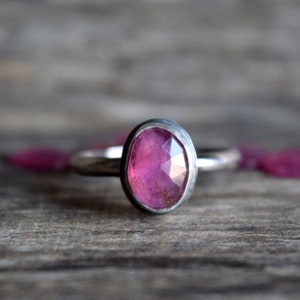 May include: A silver ring with an oval, faceted pink gemstone. The ring is set on a wooden surface, with several small pink petals scattered around it. The gemstone has a bright, reflective quality.