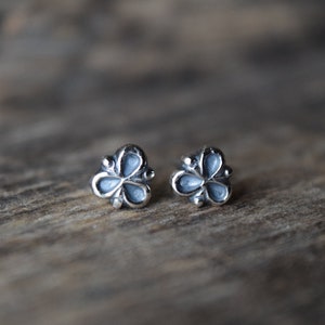May include: A pair of silver-colored stud earrings, each shaped like a three-leaf clover. The earrings have a light blue enamel filling and are set against a blurred wooden background.