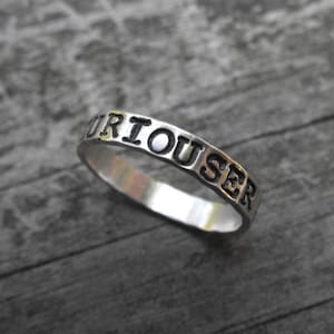 May include: A silver ring with the word "CURIOUSER" engraved on it.