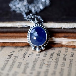 May include: A silver necklace with a blue gemstone pendant. The pendant is oval-shaped and has a faceted design. The necklace is on a stack of books.