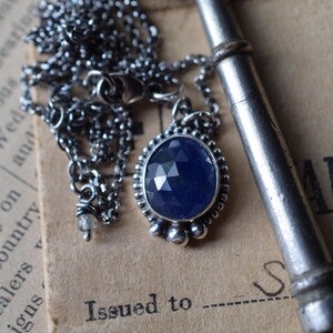 May include: A silver chain necklace with a pendant featuring a faceted blue gemstone set in a silver bezel. The pendant is surrounded by small silver beads. The necklace is lying on a piece of paper with the text "Issued to" and "Complete address" printed on it.