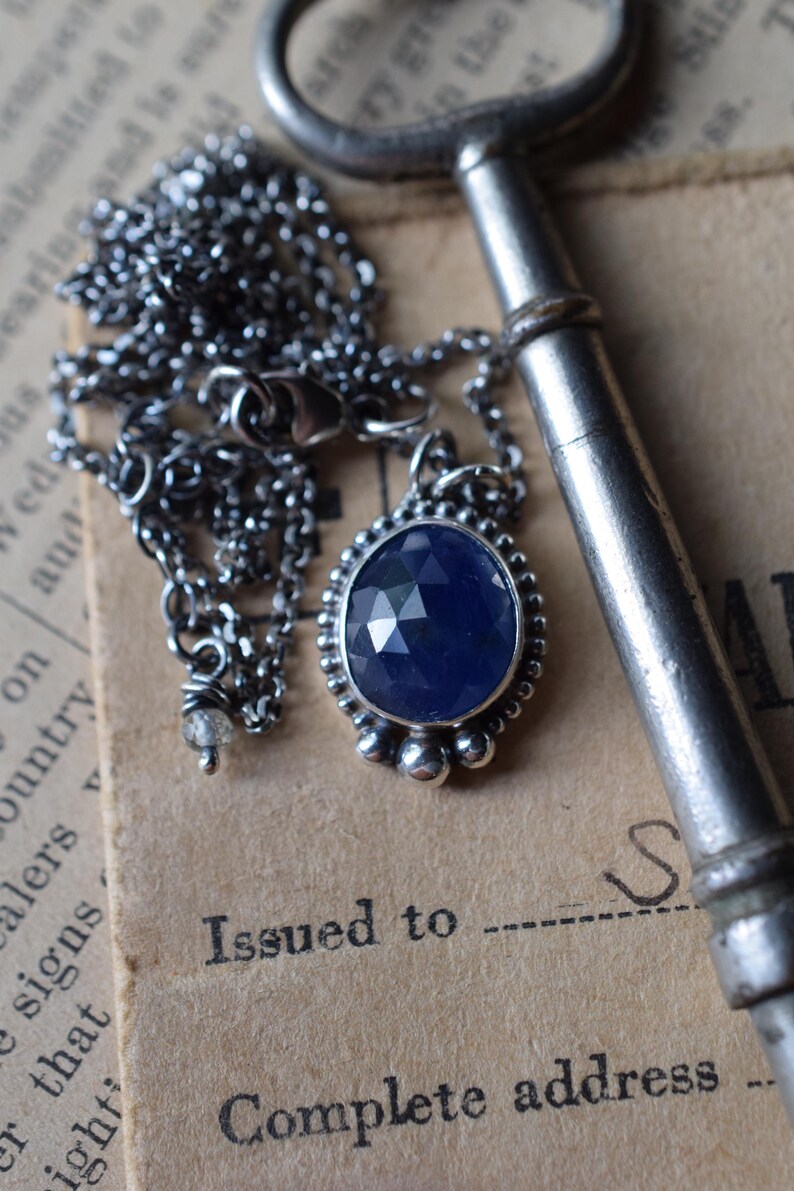 May include: A silver chain necklace with a pendant featuring a faceted blue gemstone set in a silver bezel. The pendant is surrounded by small silver beads. The necklace is lying on a piece of paper with the text "Issued to" and "Complete address" printed on it.