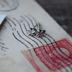 May include: A pair of silver flower stud earrings on a vintage postcard with a red stamp.