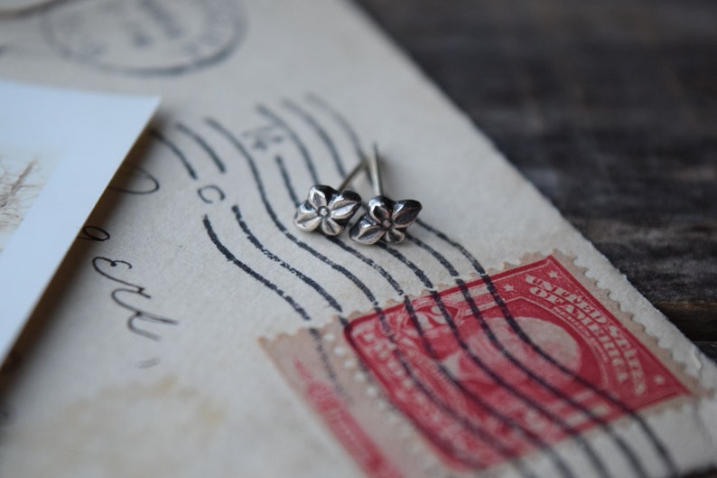 May include: A pair of silver flower stud earrings on a vintage postcard with a red stamp.