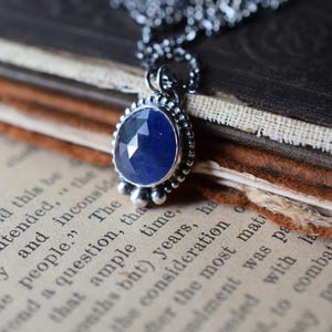 May include: A silver necklace with a blue gemstone pendant. The pendant is surrounded by small silver beads and has a faceted design. The necklace is resting on a vintage book.