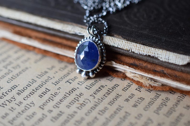 May include: A silver necklace with a blue gemstone pendant. The pendant is surrounded by small silver beads and has a faceted design. The necklace is resting on a vintage book.