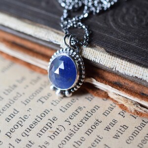 May include: A silver necklace with a blue gemstone pendant. The pendant is set in a silver bezel and has a beaded border. The necklace is lying on a book.
