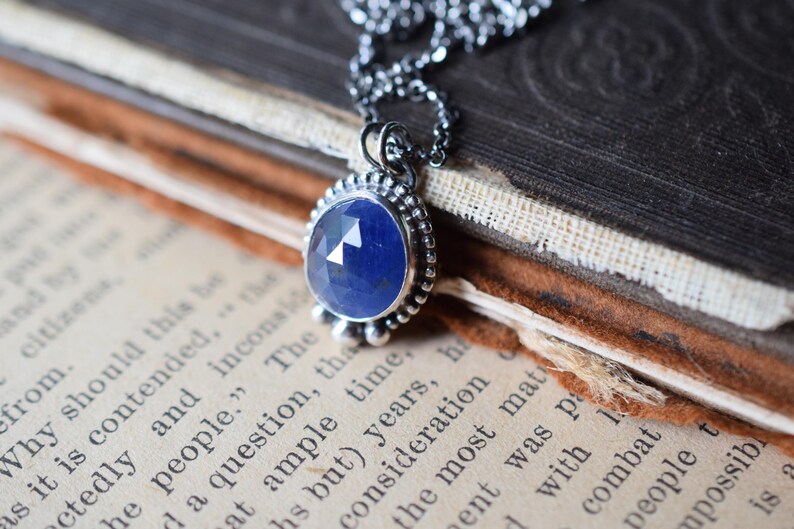 May include: A silver necklace with a blue gemstone pendant. The pendant is set in a silver bezel and has a beaded border. The necklace is lying on a book.