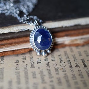 May include: A silver necklace with a blue gemstone pendant. The pendant is oval-shaped and has a faceted design. The necklace is on a stack of old books.