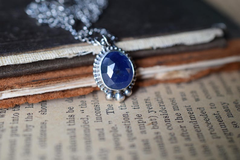 May include: A silver necklace with a blue gemstone pendant. The pendant is oval-shaped and has a faceted design. The necklace is on a stack of old books.