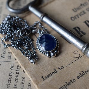 May include: A silver necklace with a dark blue gemstone pendant. The pendant is set in a silver bezel and has a textured edge. The necklace is lying on a piece of paper with text that reads "Issued to" and "Complete address".