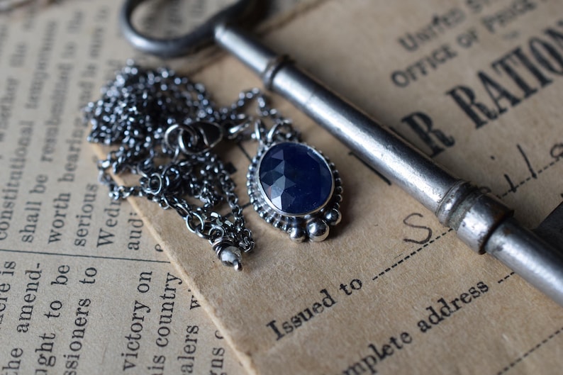 May include: A silver necklace with a dark blue gemstone pendant. The pendant is set in a silver bezel and has a textured edge. The necklace is lying on a piece of paper with text that reads "Issued to" and "Complete address".