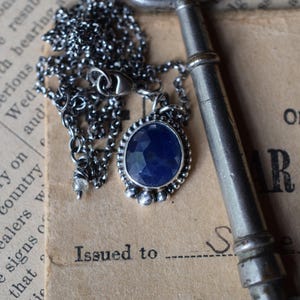 May include: A silver chain necklace with a dark blue gemstone pendant. The pendant is surrounded by small silver beads and the chain is lying on a vintage book with an old key.