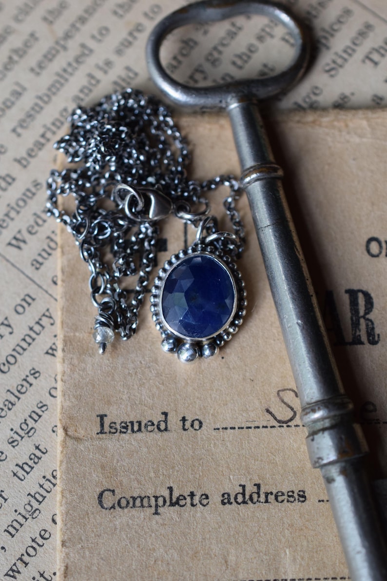 May include: A silver chain necklace with a dark blue gemstone pendant. The pendant is surrounded by small silver beads and the chain is lying on a vintage book with an old key.