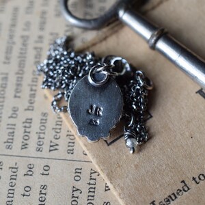 May include: A silver pendant necklace with a round, textured charm. The charm has a raised design and the initials "JR" engraved on the back. The necklace is on a silver chain.