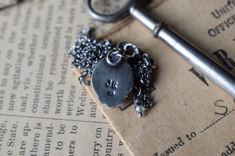 May include: A silver pendant necklace with a round, textured charm. The charm has a raised design and the initials "JR" engraved on the back. The necklace is on a silver chain.