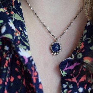 May include: A silver chain necklace with a blue gemstone pendant. The pendant is round and has a silver bezel with small silver beads around the edge.