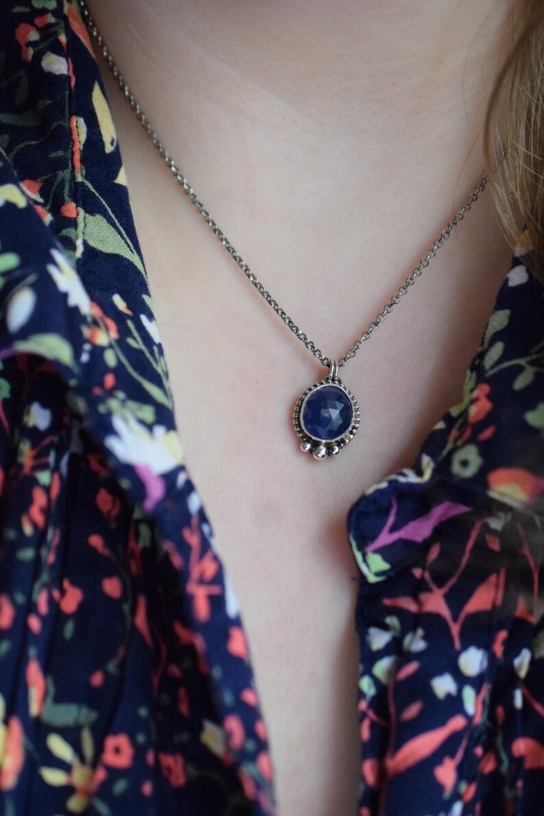 May include: A silver chain necklace with a blue gemstone pendant. The pendant is round and has a silver bezel with small silver beads around the edge.