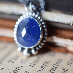 May include: A silver pendant necklace with a faceted blue sapphire gemstone. The pendant is surrounded by a silver beaded border.