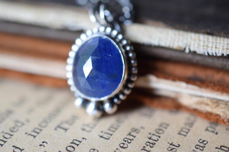 May include: A silver pendant necklace with a faceted blue sapphire gemstone. The pendant is surrounded by a silver beaded border.