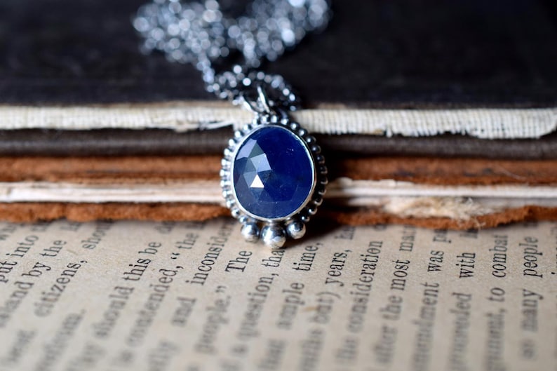 May include: A silver necklace with a blue gemstone pendant. The pendant is oval-shaped and has a faceted design. The necklace is on a stack of books.