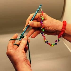 May include: A person is crocheting with a teal crochet hook and green yarn. A red coiled bracelet is on the person's wrist. A colorful chain of small, linked, plastic flowers hangs from the crochet hook.