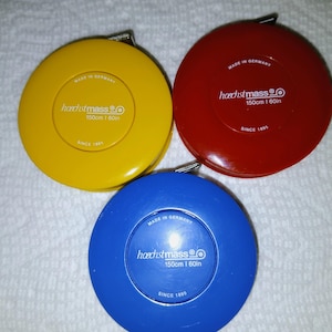 May include: Three Hoeschtmass measuring tapes in yellow, red, and blue. Each tape is 150cm (60in) long and marked "Made in Germany" and "Since 1895". The tapes are round and have a metal measuring tape pull.