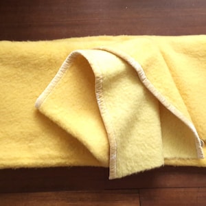 May include: A soft, bright yellow blanket with a textured, fuzzy surface and a matching yellow fabric trim along the edges. The blanket is folded over itself, showcasing the consistent solid color and the finished stitched border detail.