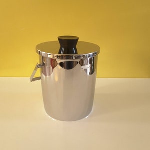 May include: A stainless steel ice bucket with a lid and handle. The bucket has a shiny, reflective surface and a black handle on the lid. The background is a solid yellow color.