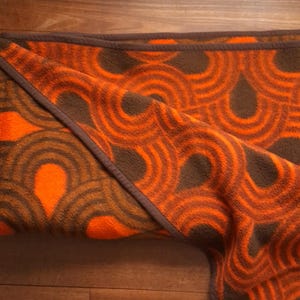 Può includere: Una coperta in pile piegata con un motivo geometrico ripetitivo di archi concentrici arancioni e marroni. Il tessuto ha una superficie morbida e testurizzata, rifinita con un bordo cucito marrone scuro lungo i lati del materiale.