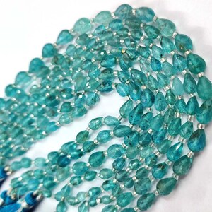 May include: A strand of teal blue gemstone beads, each bead is faceted and shaped like a teardrop. The beads are strung on a silver thread.
