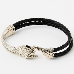 May include: A bracelet featuring a double-strand black braided cord and a silver-toned metal clasp shaped like a snake head. The snake head and tail components meet to secure the bracelet, showcasing detailed scale textures on the metal surface.
