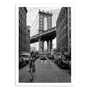 May include: A woman in a dress and jacket walks down a cobblestone street in New York City. The Brooklyn Bridge is in the background.
