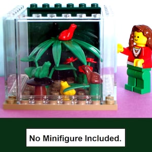 May include: A clear plastic terrarium with a Lego scene inside, featuring green plants, red flowers, and various Lego animal figures. The base is tan, and the top is dark green. The text "No Minifigure Included" is at the bottom.