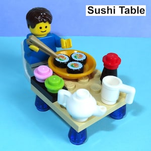 May include: A Lego sushi table set with a minifigure, sushi rolls, and condiments. The table is tan with blue legs, and the sushi is on a yellow plate. The text "Sushi Table" is at the top, and "No Minifigure!!!" is at the bottom.