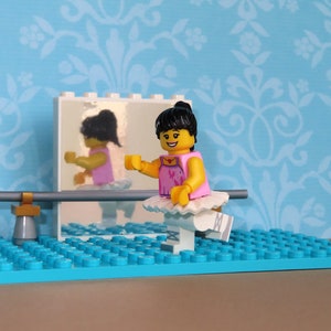 以下が含まれることがあります： ピンクのレオタードとチュチュを着たレゴのバレリーナのフィギュアが、鏡の前で練習しています。フィギュアはバレエバーのある青いベースの上にいます。背景は、花柄の薄い青色の壁です。