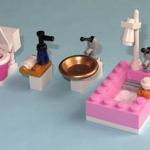 以下が含まれることがあります： カラフルなブロックで作られたミニチュアバスルームセット。 ピンクと白のトイレ、青いボトル付きのシンク、金色のボウル、白い蛇口付きのピンクのバスタブが含まれています。