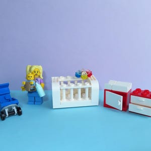 May include: A Lego scene featuring a blue stroller, a Lego figure holding a baby, a white crib, and red and white furniture. The baby is yellow and the figure has blonde hair. The crib has colorful toys on top.