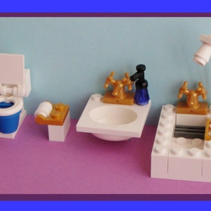 以下が含まれることがあります： 白と青のブロックで作られたミニチュアバスルームセット。トイレ、洗面台、バスタブがあります。洗面台とバスタブには金色の蛇口が付いています。白い紙のロールが小さな金色のスタンドの上に置かれています。